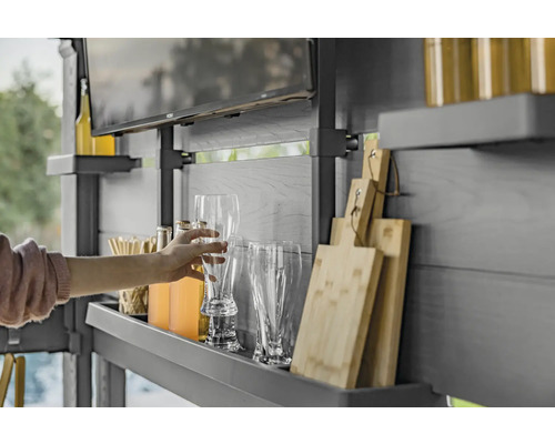 Eine Person greift nach einem Bierglas in einem Outdoor-Küchenbereich mit Regalen, Getränken, Gläsern, Schneidebrettern und einem Fernseher.