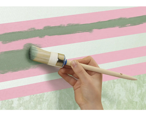 Eine Person streicht mit einem Rundpinsel eine grüne Linie auf eine Wand mit rosa und weißen Streifen