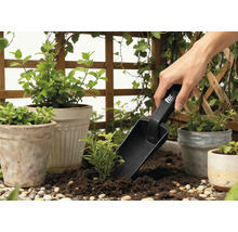 Gartenszene mit Pflanztöpfen, umgebender Begrünung und einer Person, die eine Pflanzschaufel benutzt.