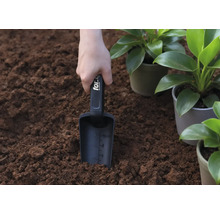 Eine Hand hält eine Pflanzschaufel in der Erde, neben eingetopften Pflanzen.