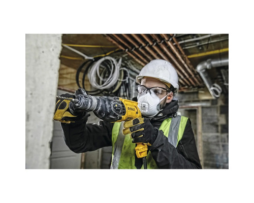 Ein Mann mit Schutzhelm, Schutzbrille und Atemschutzmaske bohrt mit einer gelben Bohrmaschine in eine Wand.