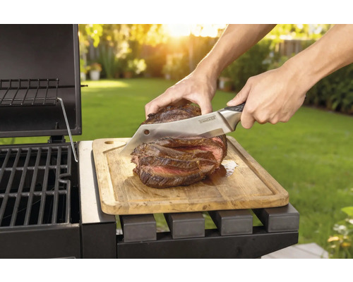 Zubereitetes Steak wird mit einem Messer auf einem Holzbrett neben einem Grill geschnitten.
