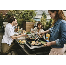 Personen grillen Gemüse auf einem Tischgrill im Freien bei einer Gartenparty.