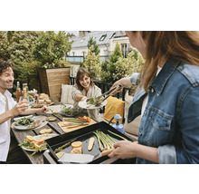 Eine Gruppe von Menschen genießt ein Mittagessen im Freien mit Grill, Gemüse und Salaten auf einem Dachgarten.