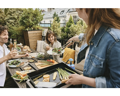 Eine Gruppe von Menschen genießt ein Mittagessen im Freien mit Grill, Gemüse und Salaten auf einem Dachgarten.