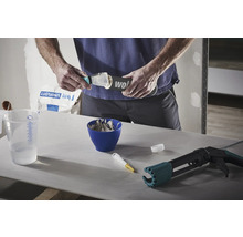 Mann füllt Spachtelmasse auf eine Spachtel, daneben befinden sich ein Messbecher, eine Schüssel mit angerührter Spachtelmasse und eine Kartuschenpistole