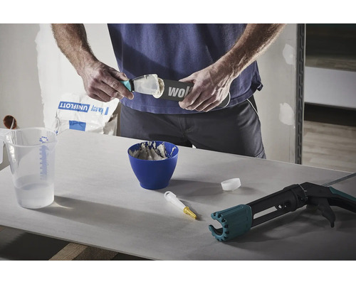Mann füllt Spachtelmasse auf eine Spachtel, daneben befinden sich ein Messbecher, eine Schüssel mit angerührter Spachtelmasse und eine Kartuschenpistole