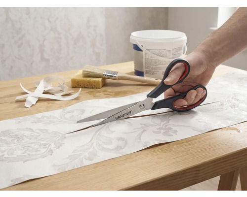 Eine Person schneidet ein Stück gemusterte Tapete mit einer Schere der Marke Rautner auf einem Tisch