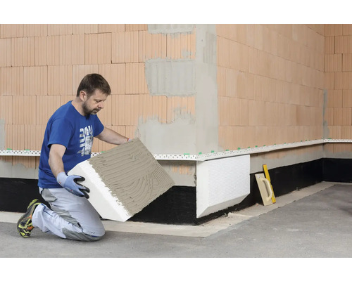 Ein Mann kniet und befestigt eine Dämmplatte an einer Wand.