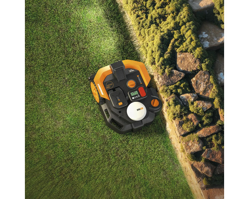 Aufsicht auf einen Rasenmähroboter auf Gras neben einer Steinmauer.