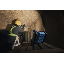 Ein Handwerker mit Schutzhelm bohrt mit einem Akkuschrauber an einer Holzdecke, während eine Arbeitsleuchte im Vordergrund steht.