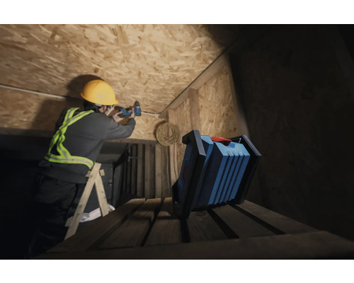 Ein Handwerker mit Schutzhelm bohrt mit einem Akkuschrauber an einer Holzdecke, während eine Arbeitsleuchte im Vordergrund steht.