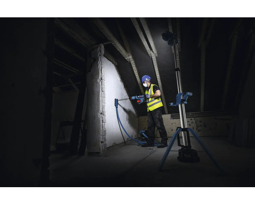 Handwerker bohrt mit Bohrhammer in eine Wand auf einem Dachboden, beleuchtet von einem Baustrahler.