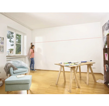 Frau benutzt einen Laser, um eine Linie an einer Wand in einem Raum zu ziehen, wo ein Tisch und ein Sessel stehen.