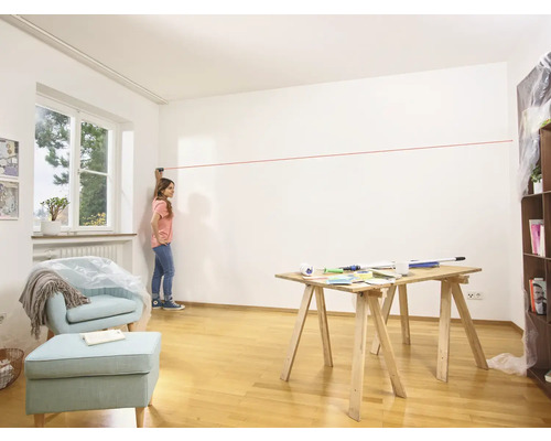 Frau benutzt einen Laser, um eine Linie an einer Wand in einem Raum zu ziehen, wo ein Tisch und ein Sessel stehen.