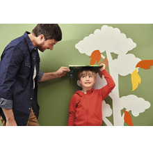 Vater misst mit einem digitalen Messgerät die Größe seines Sohnes vor einer Wand mit Baumdekoration.