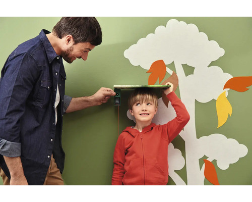 Vater misst mit einem digitalen Messgerät die Größe seines Sohnes vor einer Wand mit Baumdekoration.