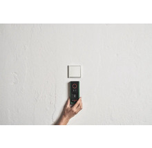 Hand hält Bosch Entfernungsmesser an einer Wand in der Nähe eines Lichtschalters.