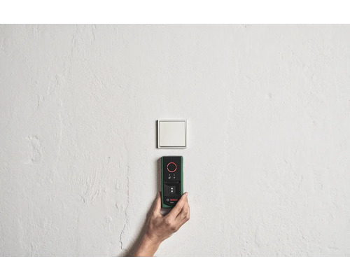 Hand hält Bosch Entfernungsmesser an einer Wand in der Nähe eines Lichtschalters.