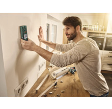 Mann verwendet ein Ortungsgerät an einer Wand in einem Raum, um Installationsarbeiten vorzubereiten.