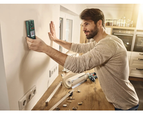 Mann verwendet ein Ortungsgerät an einer Wand in einem Raum, um Installationsarbeiten vorzubereiten.