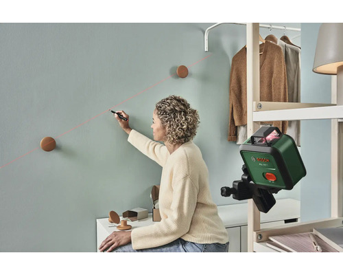 Frau markiert mit einem Laser-Nivelliergerät eine Linie an einer Wand für die Befestigung von Wandhaken