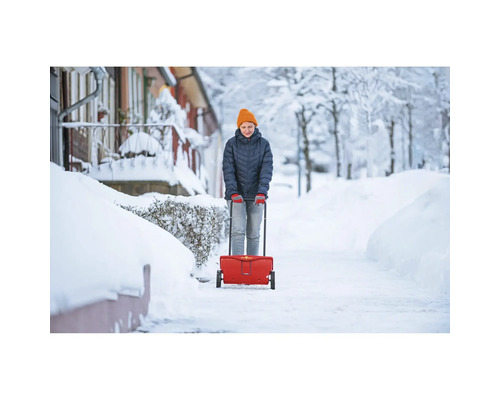 Frau benutzt einen Streuwagen für den Winterdienst auf einem verschneiten Gehweg.