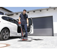 Mann saugt mit einem Akkusauger eine Fußmatte vor einem Auto.