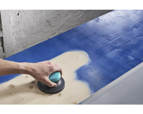 Eine Person schleift mit einem Exzenterschleifer eine blaue Holzoberfläche ab.
