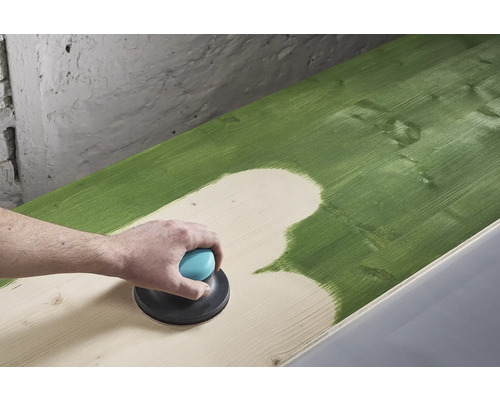 Eine Person schleift mit einem Exzenterschleifer eine grün gestrichene Holzoberfläche ab.