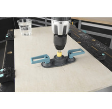 Eine Bohrmaschine mit Fliesenbohrhilfe bohrt in eine Fliese, ein Glas Wasser steht daneben.