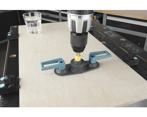 Eine Bohrmaschine mit Fliesenbohrhilfe bohrt in eine Fliese, ein Glas Wasser steht daneben.