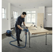 Handwerker bearbeitet eine Holzplatte mit einem Elektrohobel, der an einen Nass-Trockensauger angeschlossen ist, in einem hellen Raum.