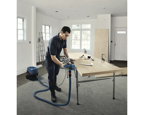 Handwerker bearbeitet eine Holzplatte mit einem Elektrohobel, der an einen Nass-Trockensauger angeschlossen ist, in einem hellen Raum.