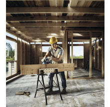 Handwerker mit Elektrohobel bearbeitet einen Holzbalken auf einer klappbaren Werkbank in einem hölzernen Rohbau.