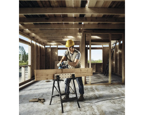 Handwerker mit Elektrohobel bearbeitet einen Holzbalken auf einer klappbaren Werkbank in einem hölzernen Rohbau.