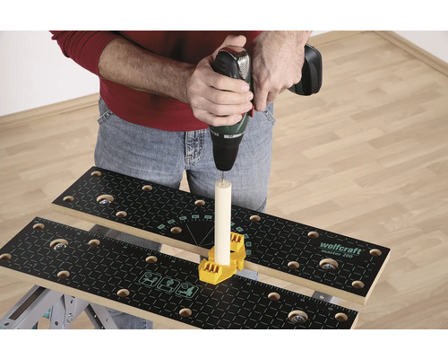 Eine Person bohrt mit einer Bohrmaschine in ein Holzstück, das auf einer Werkbank mit gelbem Befestigungselement befestigt ist.