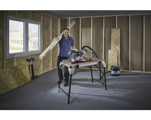 Mann mit Holzbalken, Kappsäge und Werkbank in einem Rohbau.