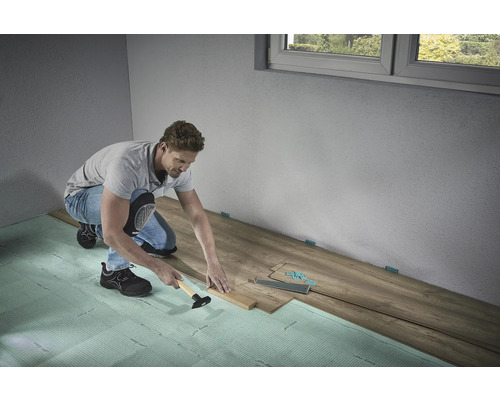 Ein Mann verlegt Laminatboden mit Hammer und Schlagholz auf Trittschalldämmung in einem Raum.