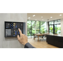 Hand bedient ein Smart-Home-Bedienpanel mit Touchscreen für Temperatur, Musik und Lichtsteuerung in einem hellen, modernen Raum.