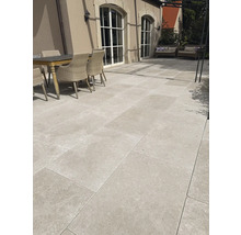 Großformatige helle Terrassenplatten in Natursteinoptik auf einer Terrasse mit Holztisch und Rattanstühlen vor einem Haus mit großen Rundbogenfenstern.