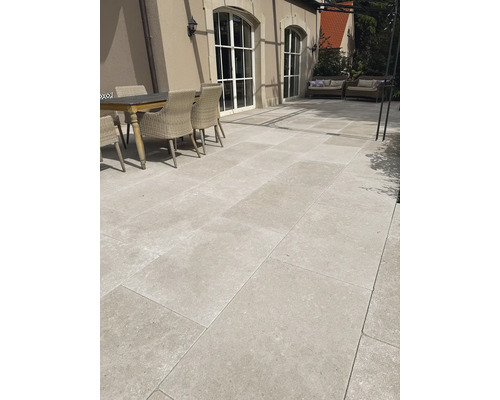 Großformatige helle Terrassenplatten in Natursteinoptik auf einer Terrasse mit Holztisch und Rattanstühlen vor einem Haus mit großen Rundbogenfenstern.