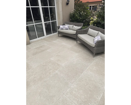 Moderne Terrasse mit hellgrauen quadratischen Terrassenplatten in Steinoptik, zwei grauen Gartensofas aus Geflecht und großen Fensterelementen.