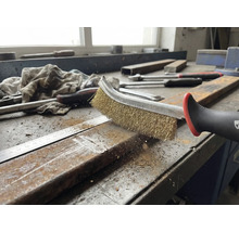 Eine Drahtbürste reinigt ein Metallstück auf einer Werkbank.