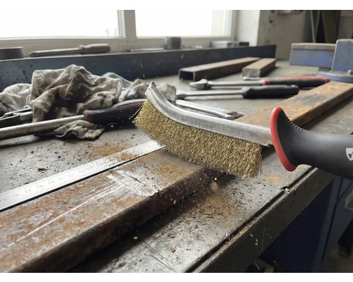 Eine Drahtbürste reinigt ein Metallstück auf einer Werkbank.