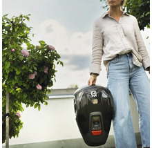 Eine Frau hält einen schwarzen Rasenmähroboter im Garten.