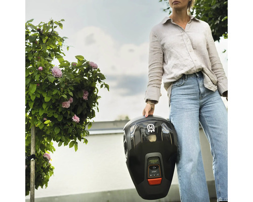 Eine Frau hält einen schwarzen Rasenmähroboter im Garten.