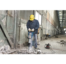 Mann mit Schutzhelm und Gehörschutz bedient einen Abbruchhammer in einem Innenraum.