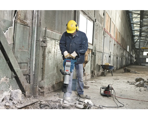 Mann mit Schutzhelm und Gehörschutz bedient einen Abbruchhammer in einem Innenraum.