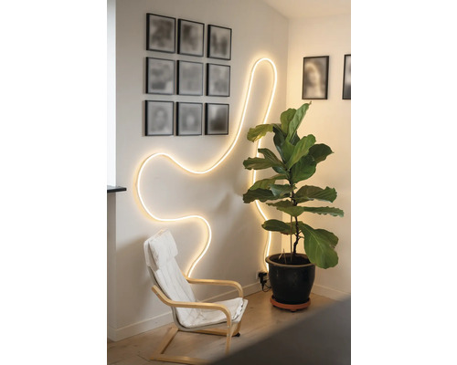 Hell beleuchteter Raum mit flexiblem LED-Lichtschlauch in Wellenform an der Wand, einem Sessel und einer großen Zimmerpflanze im Topf.